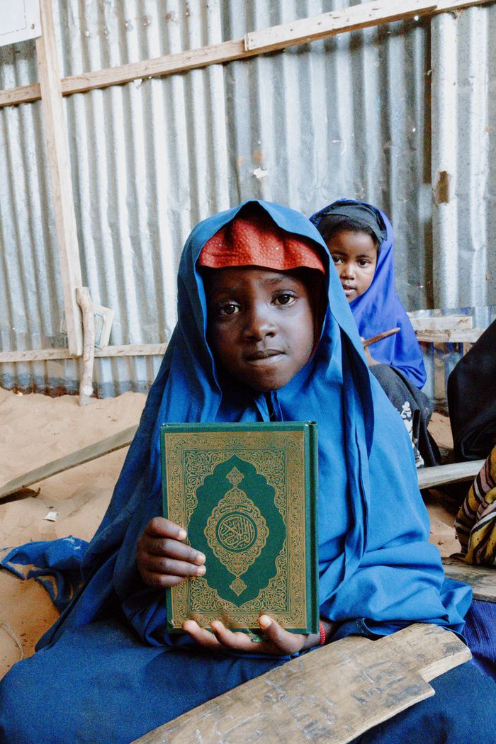 Services A child in traditional attire holding a Quran book inside a simple structure.