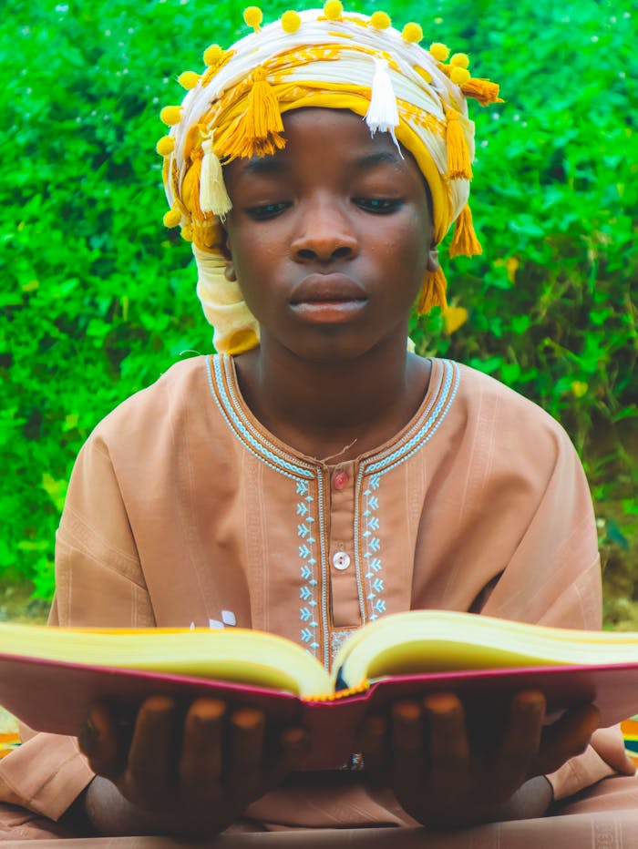 A focused African child wearing traditional clothing reads a book outdoors.